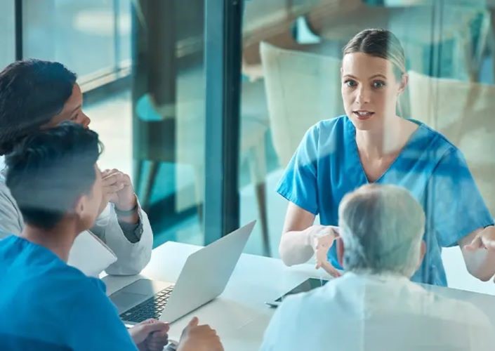 A young female nurse in blue scrubs sits at a table with three other clinicians discussing virtual care integrations and their SONIFI interactive system